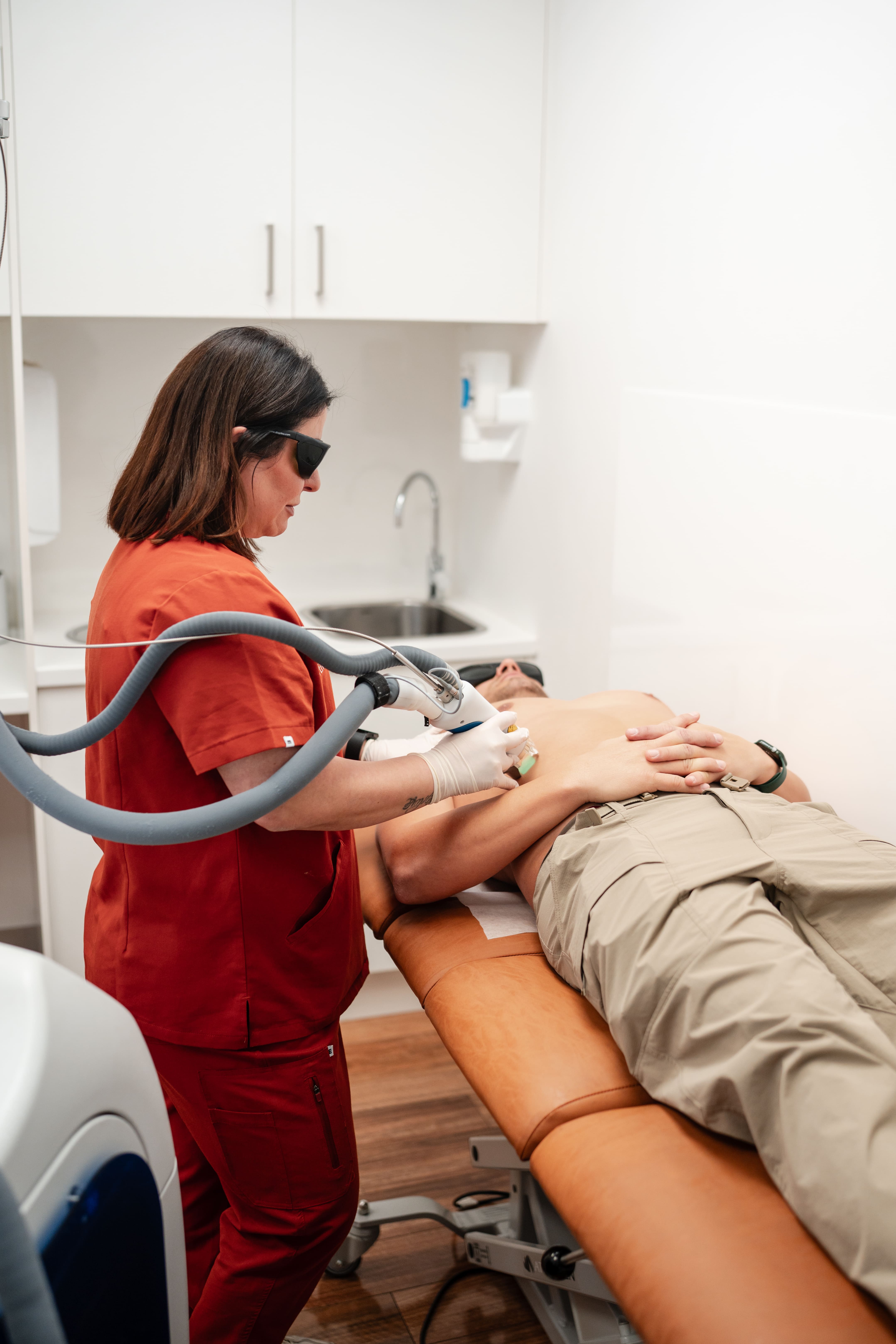 Calm, modern skin clinic treatment room with a cushioned treatment bed, soft neutral linen, warm ambient lighting and a professional clinician in a neat uniform preparing equipment